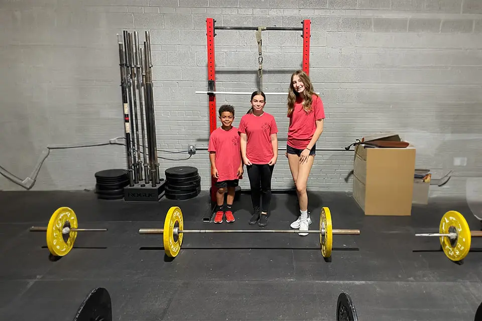 Three young people standing in front of a barbell.